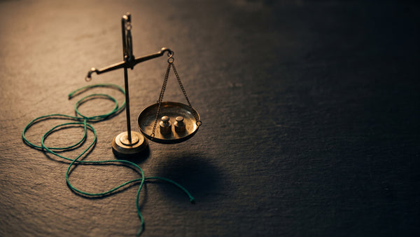 A small brass balance scale with two calibration weights on dark slate, a green linen thread alongside under warm lamp light