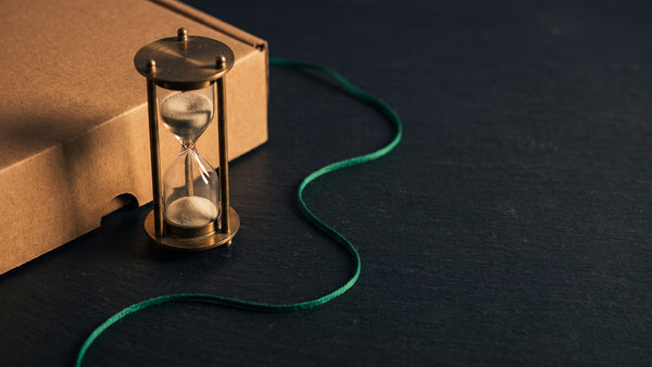 A brass hourglass beside the folded edge of a plain kraft carton on dark slate, a green linen thread curving away under warm light
